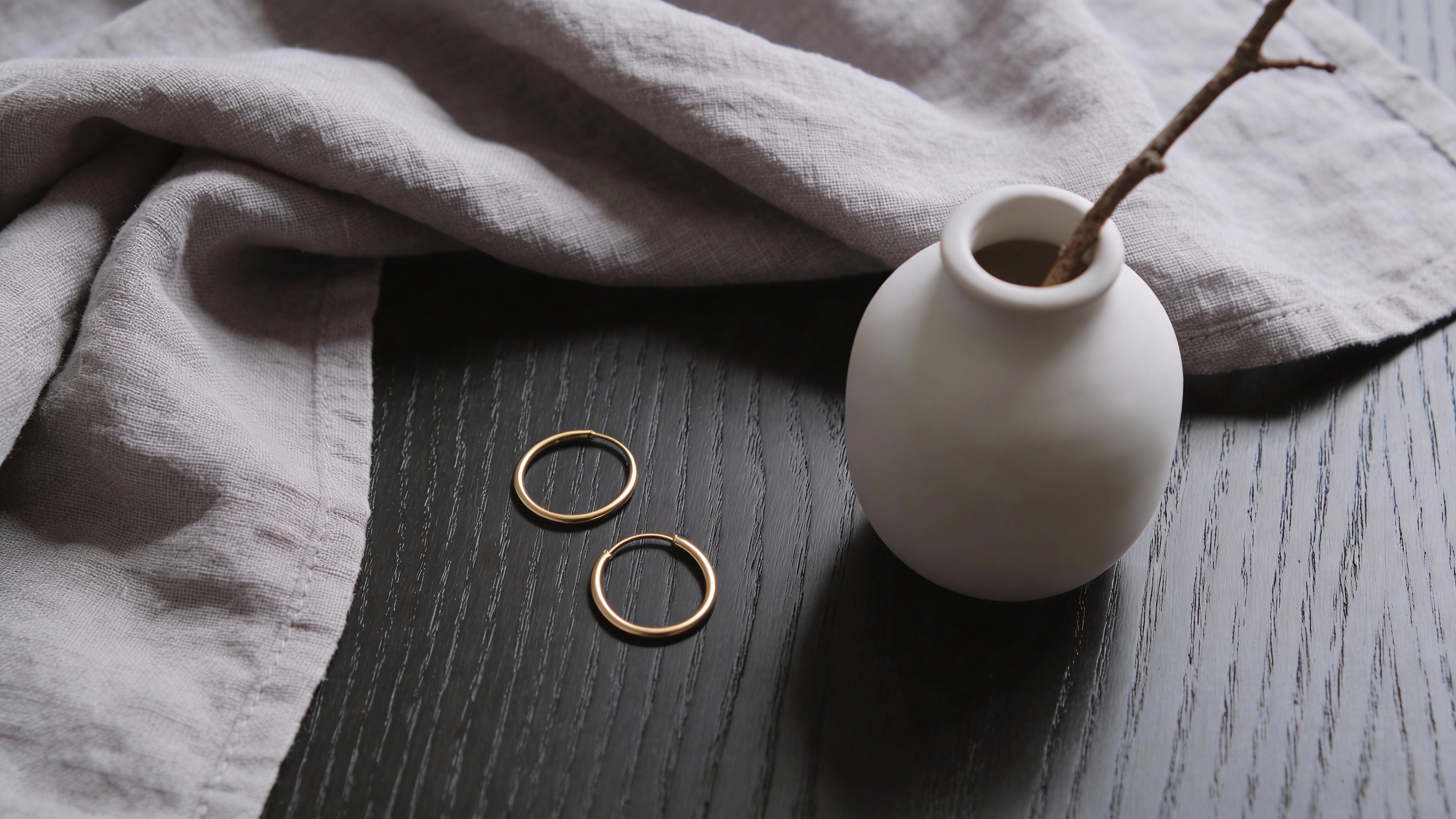 A pair of gold hoop earrings on a dark wooden surface, next to a grey linen cloth and a small white vase.