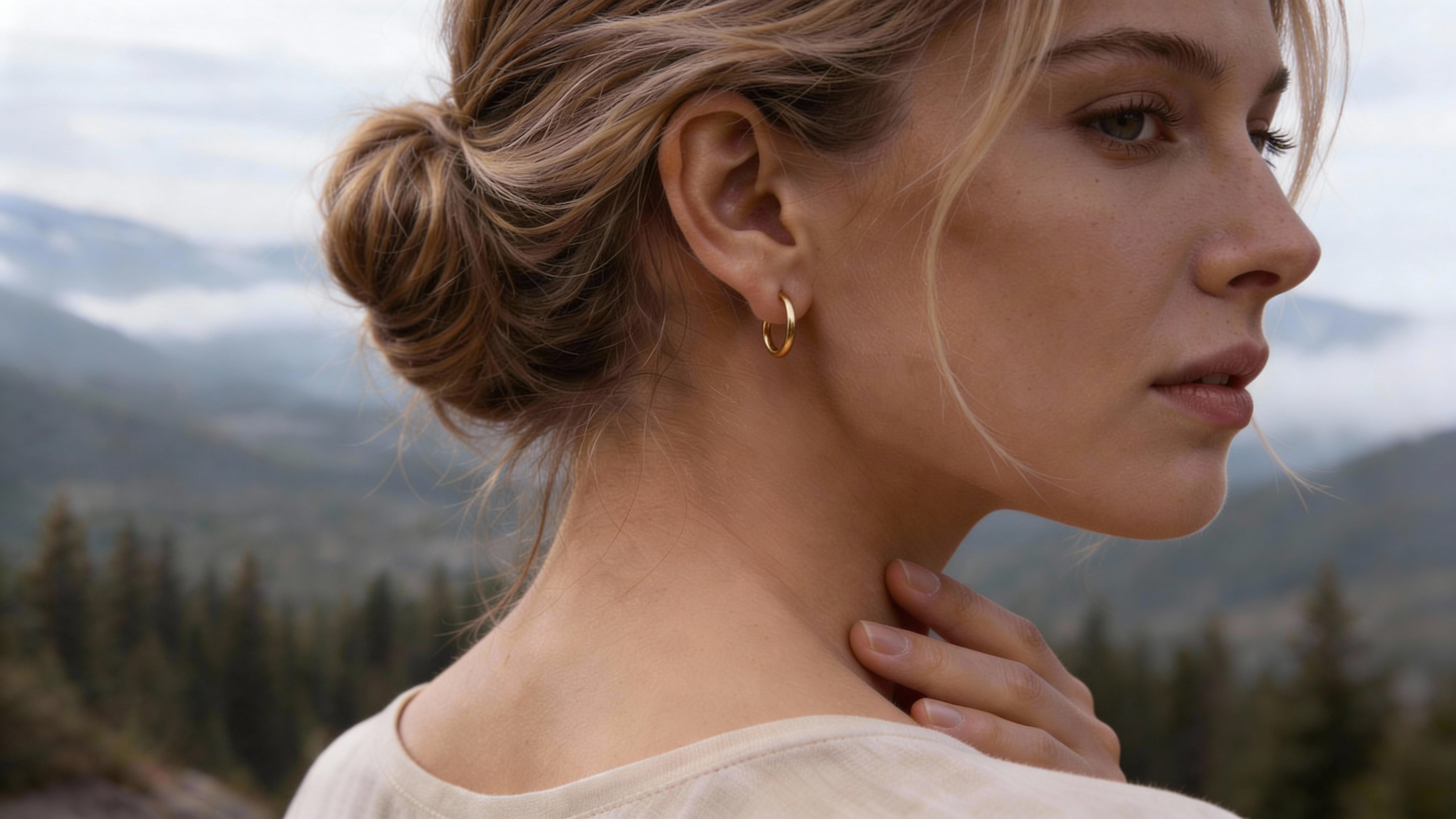 Profile of a blonde woman wearing a small gold earring against a background of Scandinavian nature.