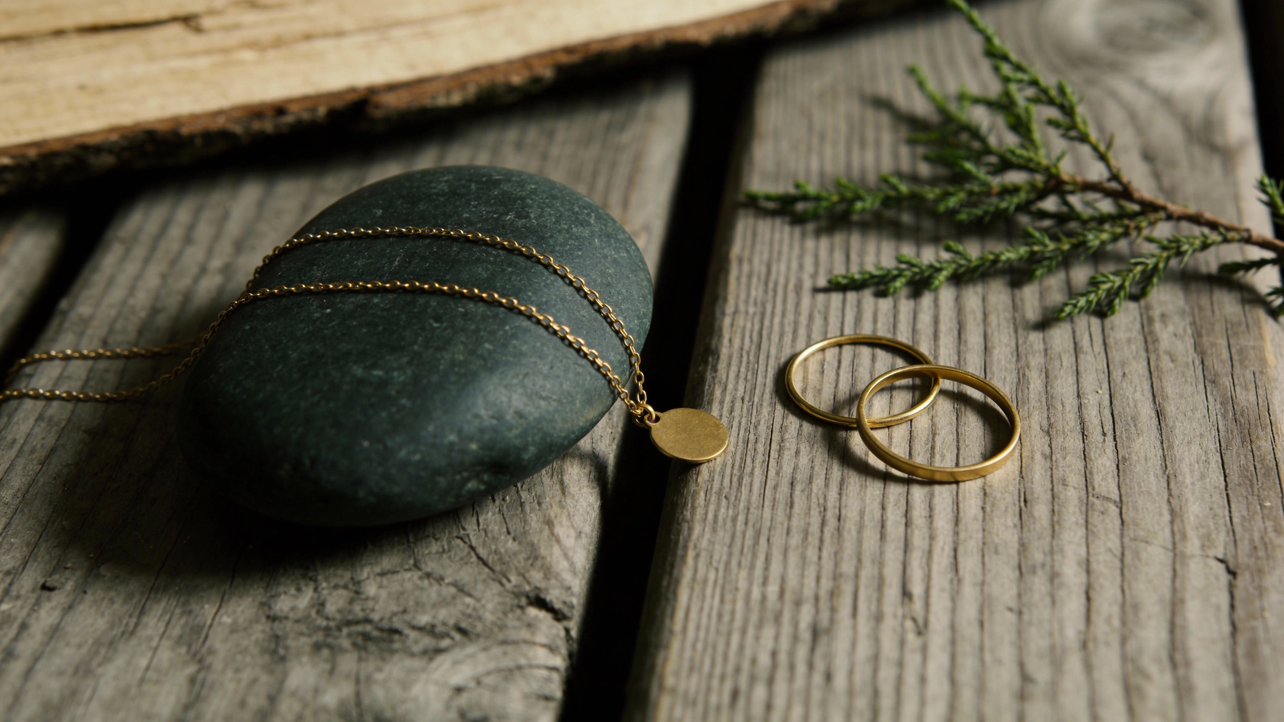 A gold anchor chain necklace with a blank coin pendant and two rings, presented on a dark stone and rustic wood planks.