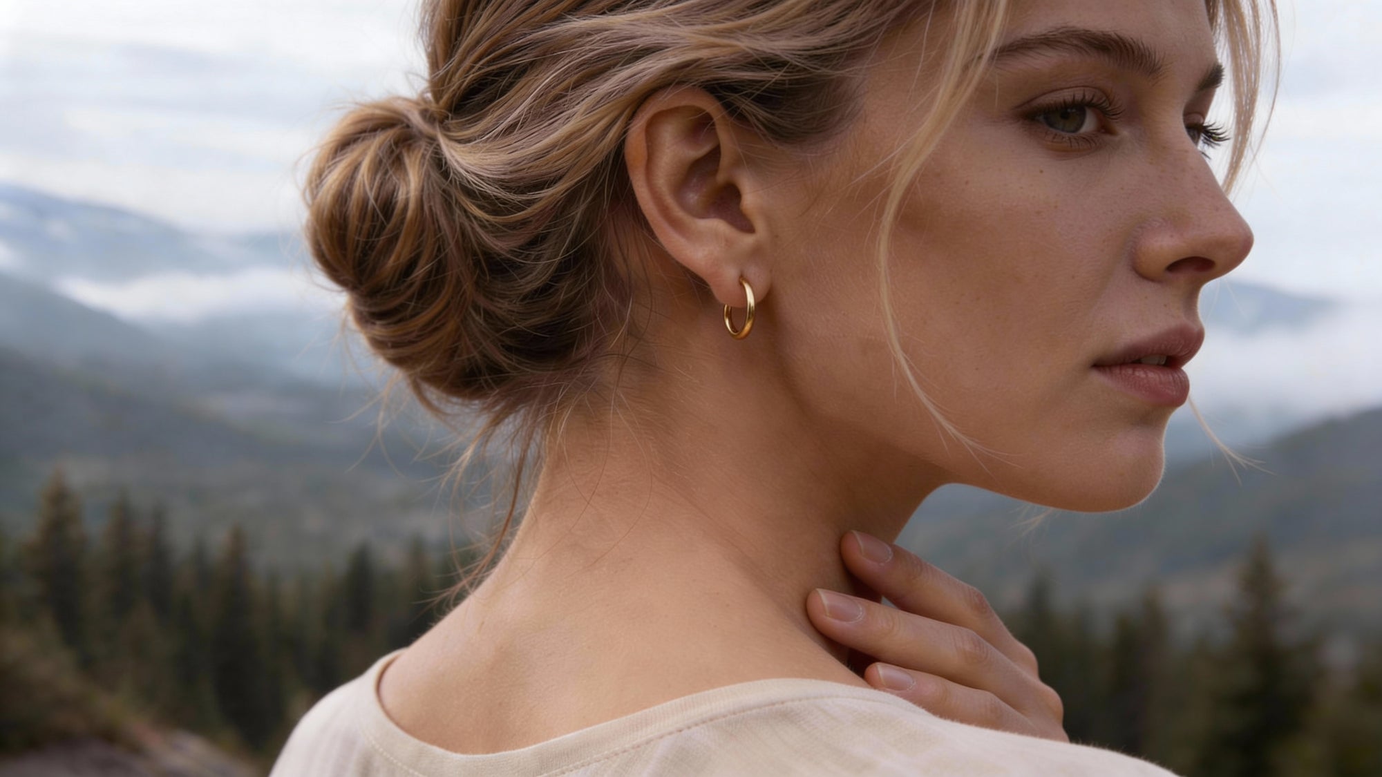 Profile of a blonde woman wearing a small gold earring against a background of Scandinavian nature.